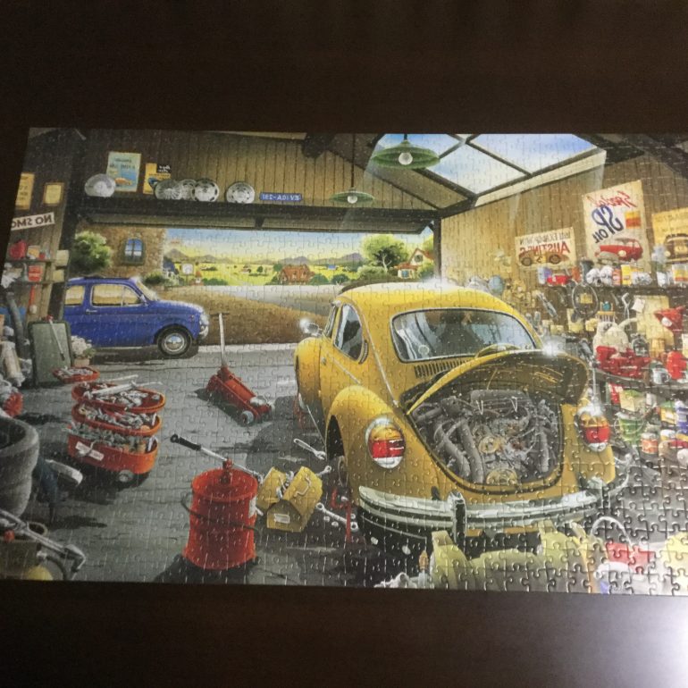 Photograph of a completed jigsaw puzzle. The puzzle shows a yellow car in a cluttered garage, a blue car in front of the garage, and a street in the distance.